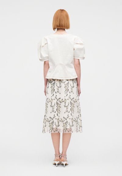 Λευκό τοπ με φουσκωτά μανίκια και στρογγυλεμένη βάση, σε συνδυασμό με φόρεμα αέρινο με κεντητά λουλούδια και διάφανο κάτω μέρος σε ανοιχτά χρώματα, φορεμένο με λευκά σανδάλια με τακούνι.