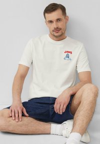 T-shirt en coton blanc à manches courtes, ornée du texte "JAWS" et d'un graphique de requin brodé en bleu et rouge, associée à un short en denim foncé.