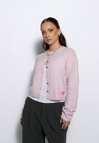 Jasnoróżowy sweter dziany z długim rękawem, zapinany na guziki, z prążkowanymi wykończeniami, noszony na białej bluzce; zdobiony małą naszywką z logo.