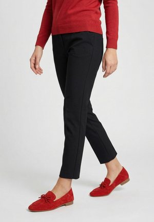 Kvinde iført korte sorte bukser, rød sweater og røde ruskindssko med lave træhæle, stående mod en ensfarvet hvid baggrund.