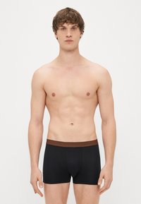 Črne bokserice z rjavim pasom, ki se ponašajo z oprijetim dizajnom in mehko teksturo. Model stoji z rokami sproščeno ob straneh pred enobarvno ozadje.