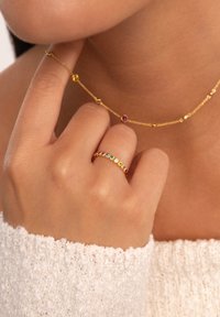 Collar de cadena de oro con piedras preciosas coloridas y un anillo de oro a juego que presenta piedras multicolores, exhibidos sobre un suéter texturizado.