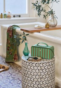 Green and brown striped cosmetic bag on a patterned black-and-white circular table. A green vase and decorative items are nearby.