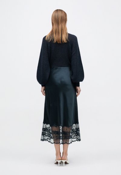 Donkere gebreide trui met pofmouwen, gecombineerd met een zwarte satijnen rok met een kanten zoom. De outfit heeft een zachte textuur en een gelaagd ontwerp.