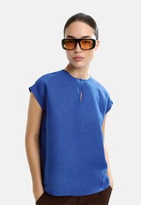 Niebieska lniana bluzka z krótkim rękawem i okrągłym dekoltem, z małym detalem w kształcie dziurki z przodu. Teksturowana tkanina, luźny krój.