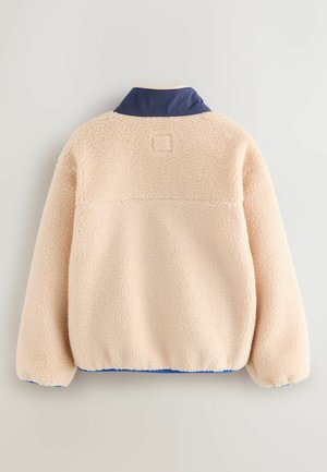 Beige fleece-takki, jossa on teksturoitu pinta, laivastonsininen kaulus ja joustavat hihansuut. Yläselän sauman yläpuolella on suorakaiteen muotoinen logo.