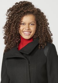 Cappotto di lana nera con colletto alto e chiusura a zip. Accenti testurizzati sulle spalle e un vivace dolcevita rosso sotto. Capelli ricci visibili.