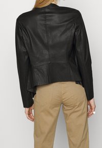 Czarna skórzana kurtka o dopasowanym kroju, gładkiej fakturze, z pojedynczym szwem z tyłu i lekko rozszerzonym dołem. W zestawieniu z beżowymi spodniami.