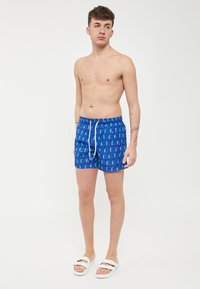 Pantaloni da bagno blu con un motivo geometrico ripetuto in colori neon. Realizzati in materiale leggero, con una vita elastica e cordoncino.