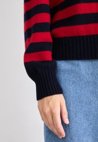 Pulover cu dungi roșii și bleumarin, cu manșete ribbate, fabricat dintr-un material tricotat moale, asortat cu blugi de denim de un albastru deschis. Prim-plan pe mânecă și mână.