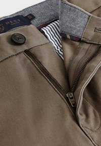 Slim-fit trousers in khaki fabric featuring a zip fly, button closure, and striped inner waistband detail. Brass zipper with YKK engraving.