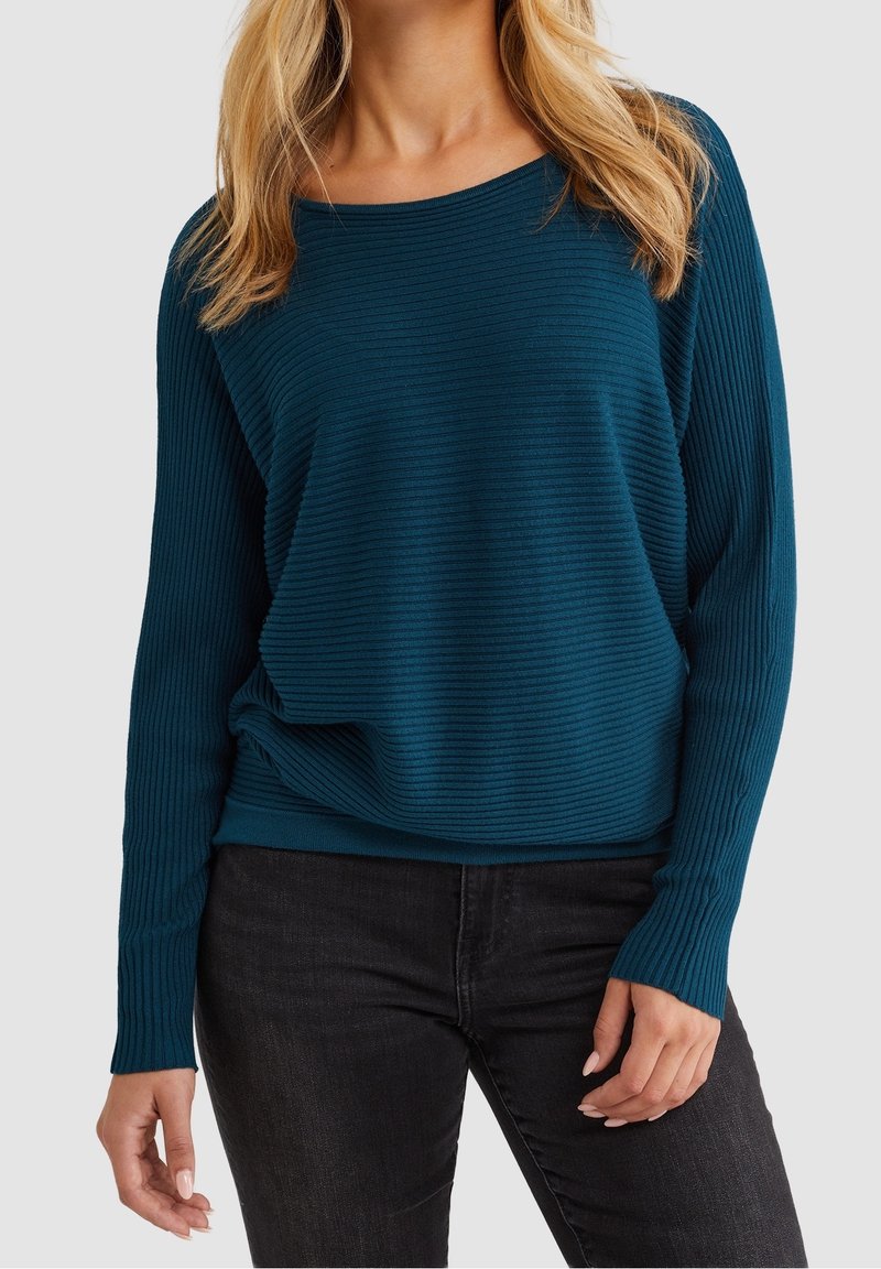Femme portant un pull côtelé à manches longues couleur sarcelle avec un jean noir, debout devant un fond uni gris clair.
