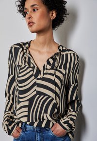 Blusa nera e beige con stampa, scollatura a V, vestibilità comoda e dettagli con colletto arricciato. Maniche lunghe con polsini aderenti. Indossata con jeans.