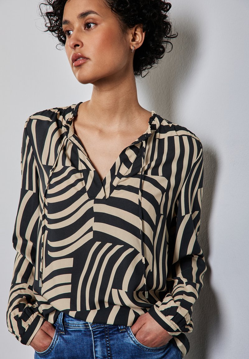 Blusa nera e beige con stampa, scollatura a V, vestibilità comoda e dettagli con colletto arricciato. Maniche lunghe con polsini aderenti. Indossata con jeans.