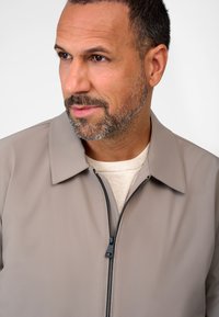 Light grey zip-up jacket with a collar, featuring a smooth texture and black zipper. Worn over a cream sweater, showcasing neutral tones.