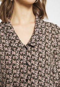Blusa floreale in tessuto marrone scuro con piccoli fiori rosa e bianchi. Presenta un collo a camicia e bottoni neri lungo la parte anteriore.