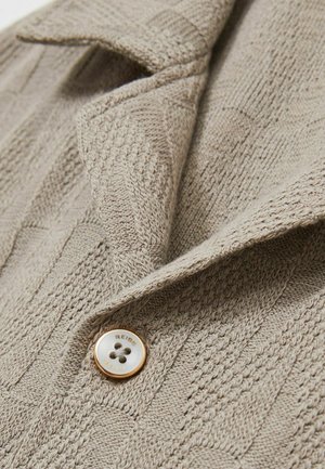 Cardigan bej tricotat, cu un model texturat, guler cu colțuri și un buton rotund cu accente aurii și un centru alb.