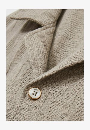 Beige kötött kardigán texturált mintázattal, kifordított gallérral és fehér középpontú, arany díszítésű gombbal.