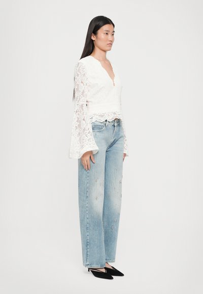Witte kant top met een V-hals en uitlopende mouwen, gecombineerd met lichtblauwe distressed jeans met versieringen en zwarte puntige schoenen.