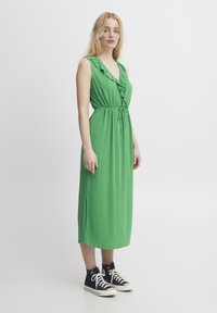 Rochie verde fără mâneci, cu decolteu volănaș, talie cu șnur și fante laterale. Purată cu adidași negri cu vârf înalt. Textură netedă.