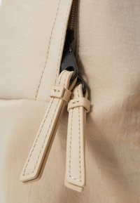 Zaino di tessuto beige con una texture liscia, chiusura con zip dotata di tiranti in pelle beige e accessori neri.