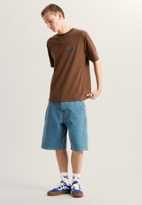 Bruine katoenen T-shirt met een kleine groene en blauwe graphic op de linkerkant van de borst, gecombineerd met lichtblauwe denimshorts en blauwe sneakers.