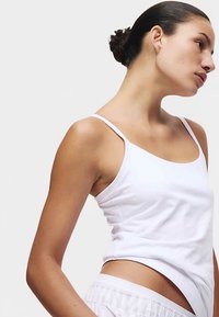 Weißes Baumwolltanktop mit dünnen Trägern, das eine taillierte Passform und eine glatte Textur aufweist. Kombiniert mit lockeren, hellfarbenen Shorts.