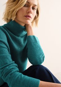 Maglione a collo alto teal con texture a coste, maniche lunghe e polsini aderenti, indossato con pantaloni blu scuro. Modella che posa con una mano sul collo.