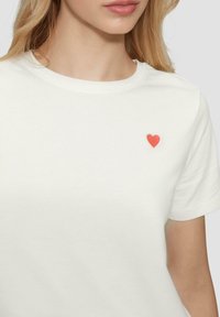 Witte katoenen t-shirt met een klein rood hartje geborduurd op de linkerborst, met een klassieke ronde hals en korte mouwen.