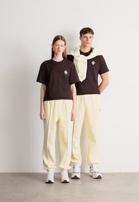 Δύο μοντέλα φοράνε ταιριαστά σύνολα: μια μαύρη μπλούζα με γεωμετρικό λογότυπο, εκρού φόρμες και λευκά αθλητικά παπούτσια. Απλό, άνετο φόρεμα.