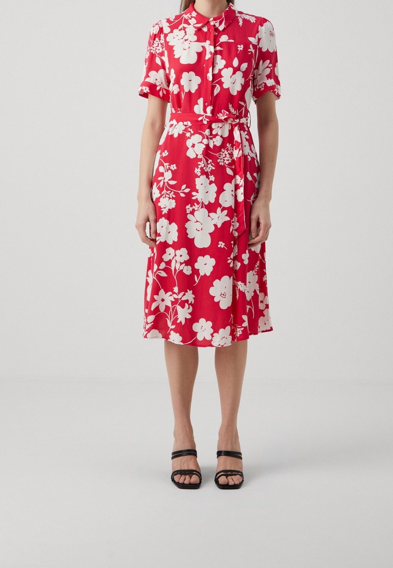 Femme portant une robe-chemise midi rouge avec imprimé floral blanc, manches courtes, ceinture à la taille et sandales à talons noires à bout ouvert.