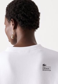 White cotton t-shirt with a round neckline and short sleeves, featuring a black printed label on the back with Lacoste branding.