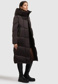 Pitkä tummanruskea toppatakki, jossa on korkea kaulus, tikattu kuvio, vetoketjullinen sivutasku ja sileä pinta.