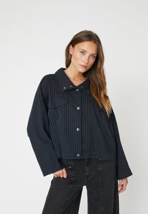 Navy blue cropped jacket with white vertical stripes, large collar, front buttons, and two chest pockets. Worn with dark jeans.