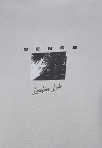 Tela gris con el texto negro "SENSE" encima de un cuadrado abstracto en blanco y negro y "Lossless Lab" escrito a mano debajo.