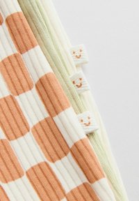 Pehmeä ribbineulospinta, jossa oranssin ja valkoisen ruutukuvio, vierekkäin vaaleanvihreän kankaan kanssa, molemmissa pienet valkoiset liput, joissa oranssit hymynaamat.