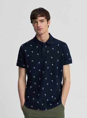 Młody mężczyzna ubrany w granatową koszulkę polo z małymi białymi wzorami palm, stoi z rękami w kieszeniach zielonych spodni na jednolitym tle.