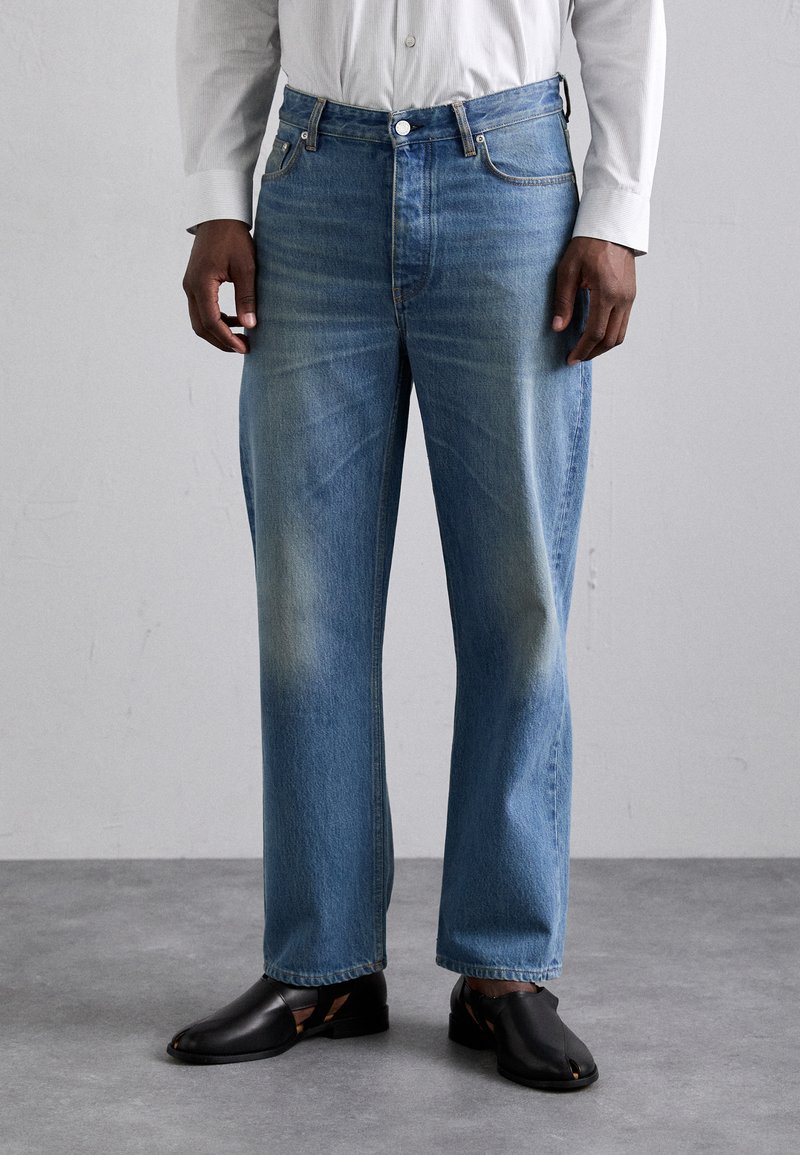 Jeans de mezclilla azul claro con un corte relajado, que presentan detalles desgastados y un diseño de cinco bolsillos, combinados con zapatos negros de punta abierta.