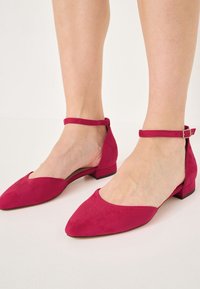 Stopy w różowych zamszowych butach na niskim obcasie z paskiem w kostce i ostrym noskiem na jasnym podłożu.