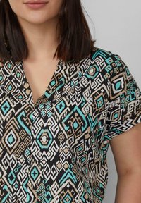 Korte mouwen blouse met een geometrisch patroon in zwart, turquoise en beige, met een V-hals en een lichtgewicht, gestructureerde stof.