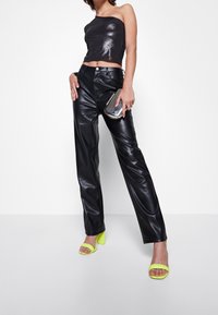 Frau in glänzenden schwarzen Lederhosen, einem schwarzen Glitzer-Top mit einem Schulterträger, neon-gelben Blockabsatzsandalen und einer metallisch-silbernen Clutch.