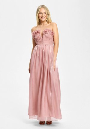 Uśmiechnięta kobieta w różowej, długiej do ziemi sukni bez ramiączek z plisowanym materiałem i kwiatowymi zdobieniami na gorsecie, w sandałach na odkryte palce.