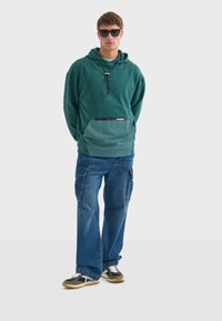 Sudadera verde con un bolsillo en contraste color teal, acentos negros y cordón. Combinada con jeans cargo azules de corte holgado y zapatillas negras.