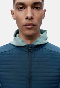 Tamsiai mėlyna zip-up striukė su tekstūruotu audiniu, kuriame yra šviesiai mėlynas gobtovas ir raukšlėtos detalės. Priekinis užtrauktukas ir lygus paviršius.