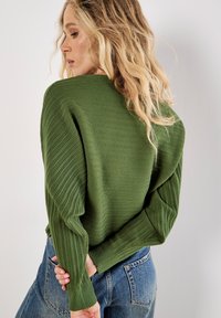 Maglione verde a coste con maniche lunghe, caratterizzato da un tessuto testurizzato e una vestibilità comoda. Indossato con jeans di denim a vita alta.