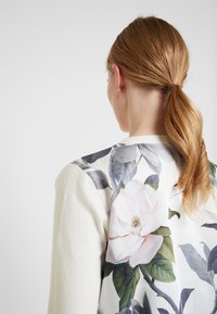 Žena s červenými vlasy v ohonu má na sobě bílé tričko s velkým květinovým potiskem růžové a bílé květiny se zelenými listy na zádech.