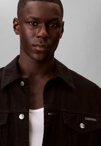 Joven hombre negro con cabello corto, vistiendo una chaqueta negra de Calvin Klein sobre una camiseta sin mangas blanca acanalada, mirando al frente con una expresión neutral.