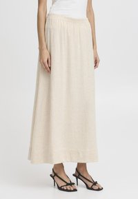 Jupe maxi beige avec une texture apparente, taille élastique et une silhouette fluide, associée à des sandales à talons fins noires.