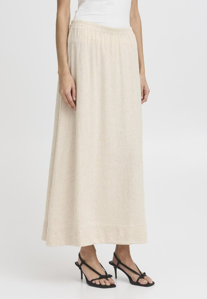 Jupe maxi beige avec une texture apparente, taille élastique et une silhouette fluide, associée à des sandales à talons fins noires.