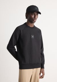 Μαύρη φόρμα με στρογγυλή λαιμόκοψη και ραγλανισμένα μανίκια, διακοσμημένη με ένα μικρό μαύρο τετράγωνο λογότυπο στο στήθος. Λείο υλικό, συνδυασμένο με μπεζ παντελόνι.